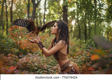 Fantasy woman with falcon. wild girl, wanders in jungle hold tamed bird. Art Photography. Amazon in  brown seductive, leather suit costume. Hairstyle dreadlocks. Autumn forest yellow, red leaves trees