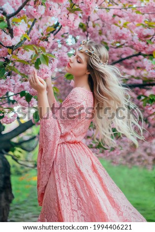Fantasy woman princess walks in spring blooming garden. hand touches pink flowers sakura tree summer nature. long lace dress wide sleeves. Blonde hair flies in wind. Elf diadem tiara crown on head