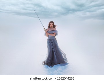 Fantasy Woman Princess Warrior Holding Medieval Iron Sword In Hands. Fairy Tale Snow Queen. Vintage Gray Blue Dress. White Winter Nature Background, Valley, Tree. Elf Girl With Steel Blade. Art Photo