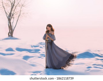 Fantasy Woman Princess Warrior Holding Medieval Iron Sword In Hands. Fairy Tale Snow Queen. Vintage Gray Blue Dress. White Winter Nature Background, Valley, Tree. Elf Girl With Steel Blade. Art Photo