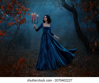 Fantasy woman queen runs in dark forest, hands hold vintage burning candlestick three candles. Blue velvet long medieval dress fly in wind. Gothic girl princess. Autumn nature night bare trees fog art