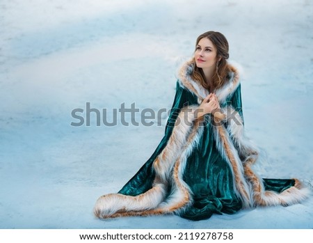 Fantasy woman sits on white snow in winter forest. Princess girl smiling face. Green long velvet vintage coat fur. Mystical image of frozen lady wanderer. Nature frost cold. Fairy tale art photography