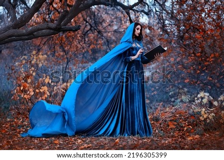 Fantasy Woman witch holds magic book in hands reads spell. Blue vintage clothes silk cape dress flies in wind. Mystery girl elf in hood. Nature autumn forest trees orange leaves. Holiday halloween