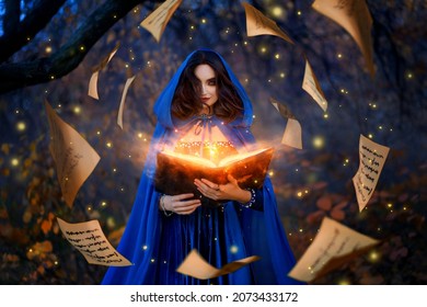 Fantasy woman witch magician in hood holds in hands magic book, bright orange light spells, wind scatters fall sheets paper page levitation. Girl sorceress. Medieval cloak blue dress magician costume - Shutterstock ID 2073433172