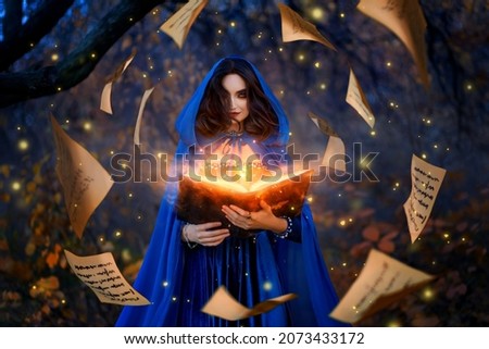 Fantasy woman witch magician in hood holds in hands magic book, bright orange light spells, wind scatters fall sheets paper page levitation. Girl sorceress. Medieval cloak blue dress magician costume
