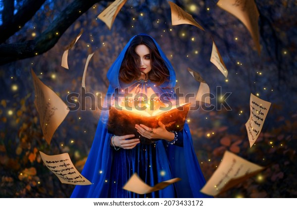 Fantasy woman witch magician in hood holds in\
hands magic book, bright orange light spells, wind scatters fall\
sheets paper page levitation. Girl sorceress. Medieval cloak blue\
dress magician costume