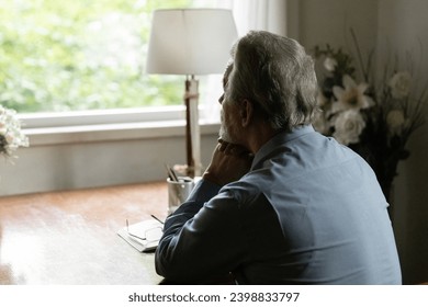 Far away in thoughts. Rear view of aged hoary male sit by table at home alone look at window ponder prop chin on folded hands. Thoughtful mature man going down to memories reviewing past. Copy space - Powered by Shutterstock