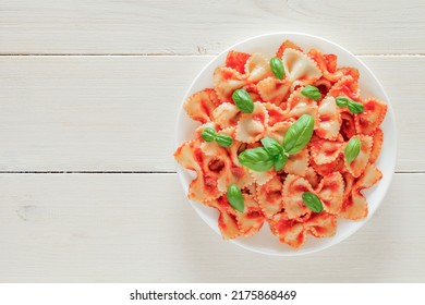 Farfalle, cooked pasta Italiano with tomato sauce and basil green leaves, in plate, on white wooden board background, top view, space to copy text.