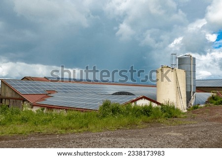 Farm buildings with rooves covered with solar panels and grain silos under a cloudy sky; view of solar panels on the rooves of barns and farm buildings
