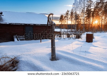 Farm pump in the snow with the sun setting behind the trees in the background