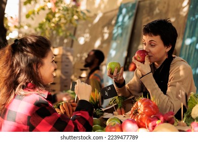 Farm to table. Female farmers market vendor smelling red apple while selling fresh organic fruits and vegetables to customer on sunny autumn day. Young happy woman buying locally grown healthy food - Powered by Shutterstock