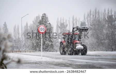 A farm tractor driving past 100km road sign in the heavy snowfall. Ashburton Lakes. South Island. 