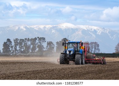 A Farm Worker In A Blue Tractor Seeding A Field In Canterbury, New Zealand