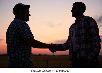 Farmer and agronomist silhouettes shaking hands standing in a wheat field after agreement in dusk. Agriculture business contract concept. Combine harvester driver and rancher handshake. Negotiations.
