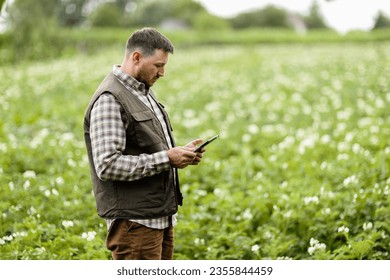 Farmer or agronomist uses digital tablet to analyse and check the growth and disease of the blooming plants in the potato field. Smart farming technology and agriculture business concept - Shutterstock ID 2355844459