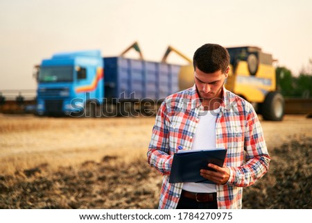 Farmer controls loading wheat from harvester to grain truck. Driver holding clipboard, keeping notes, cargo counting. Forwarder fills in consignment waybills. Agricultural commodities logistics.