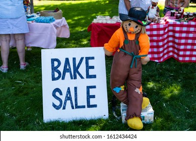 Farmer Doll Holds Bake Sale Sign At Outdoors. Different Bakery Products And Sweets On Table. Bakery Fair In Okanagan Valley, British Columbia, Canada