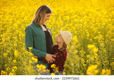 Farmer Family Mother And Daughter Embracing In Summer Shirt Laughing And Smiling At Sunset, Side View Kid And Woman In The Flowering Rapeseed Field,