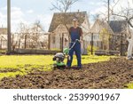 A farmer in the garden tills the land with a motorized cultivator or power tiller, preparing the soil for planting crops.
