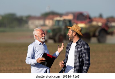 Farmer And Insurance Sales Rep Representative Discussing Agreeing Laughing In Front Of Tractor. Two Men Talking In Cultivated Field Satisfied.