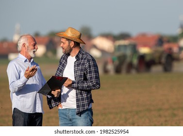 Farmer And Insurance Sales Rep Representative Talking, Discussing. Two Men Standing In Field Negotiating Holding Clipboard In Front Of Tractor Farming Machinery