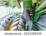 Farmer Repairing Hydraulic Hose Under Combine Undercarriage, Wearing Yellow Gloves, Using Wrenches, Propped by Dual Tires, Agricultural Equipment Maintenance, Combine Harvester Repair sstkIndustry