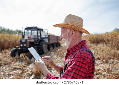 A farmer stands in a field using a tablet while a tractor is parked nearby. The day is clear, and the crops are ready for harvest.