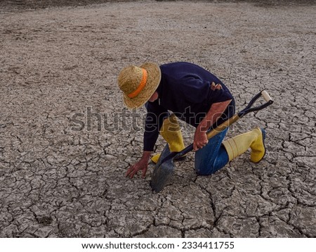 Farmer with a straw hat and a shovel is kneeling touching the dry, arid soil. Praying for rain. Drought concept.