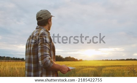 Farmer with a tablet computer in front of a sunset agricultural landscape. Man in a countryside field. Country life, food production, farming and technology.