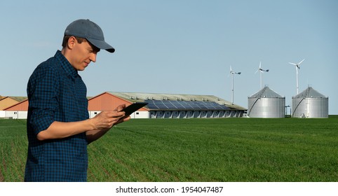 Farmer With Tablet Computer On A Background Of Modern Dairy Farm Using Renewable Energy, Solar Panels And Wind Turbines