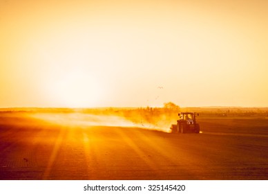 A farmer in a tractor prepares his field as the sun begins to set. The tractor is backlit by the setting sun. The sun is setting behind a low row of hills in the far distance.