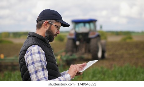 Farmer Uses A Specialized App On A Digital Tablet PC On The Background Of Working Tractor With A Cultivator In The Field