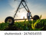Farmer is using a mobile phone while standing in a corn field at sunset near a center pivot irrigation system, holding a tablet and wearing casual clothing and boots