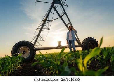 Farmer is using a mobile phone while standing in a corn field at sunset near a center pivot irrigation system, holding a tablet and wearing casual clothing and boots