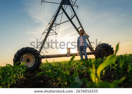 Farmer is using a mobile phone while standing in a corn field at sunset near a center pivot irrigation system, holding a tablet and wearing casual clothing and boots
