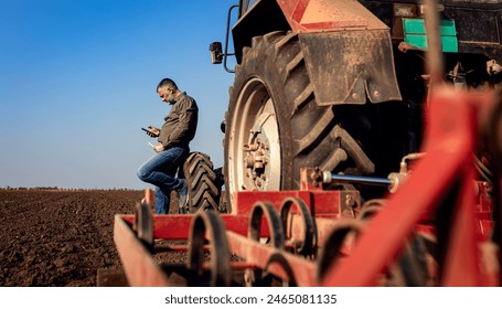 Farmer using smart phone while resting after cultivation of soil leaning on tractor tire.