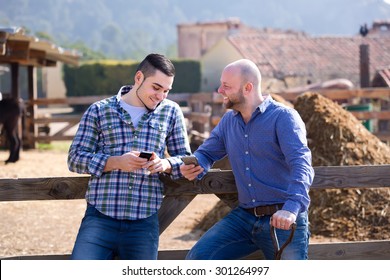 Farmers Resting After Labour Outdoors. Two Men Leaning On A Fence And Browsing Their Phones