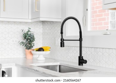 A farmhouse kitchen sink detail shot with a black faucet, mosaic tile backsplash, marble countertops, and white cabinets. - Shutterstock ID 2160564267
