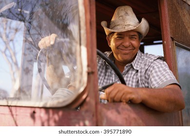 Farming And Cultivations In Latin America. Portrait Of Middle Aged Hispanic Farmer Sitting Proud In His Tractor At Sunset, Holding The Steering Wheel. He Looks At The Camera And Smiles Happy.