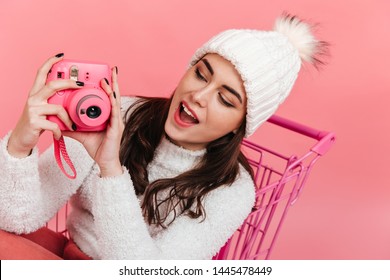 Fascinated Brunette In Knitted Hat Makes Photo On Camera Instax. Portrait Of Young Girl In Pink Trolley On Isolated Background