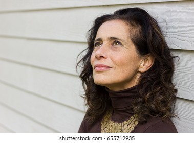 Fascinating mature woman with optimistic look leans on white wood plank wall background. Classy middle aged lady with hopeful, positive expression. Plan, imagination, creative concept