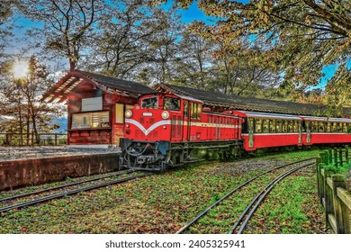 Fascinating red forest train slowly passing by the scenic platform at Dueigaoyue Station within the Alishan National Forest Recreation Area in Taiwan. This famous alpine railway features an attractive arc track bending gracefully through lush green trees and natural woodland. The iconic red locomotive and vintage carriages create a beautiful contrast against the dense foliage and mountain forest. Perfect for travel concepts, historical railway heritage, and Taiwanese tourism promotions, this picturesque landscape captures the nostalgic charm, misty atmosphere, and tranquil beauty of Alishan's high-altitude rail adventure during a bright morning.