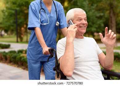 A Fascinating Talk On The Phone. Happy Old Man Sitting In A Wheelchair And Talking On The Phone, While A Nurse Rolls It Over A Sunny Park