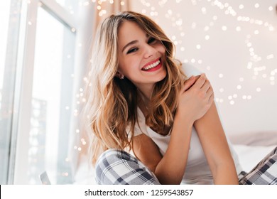 Fascinating white girl with light makeup posing in her room. Glad young woman sitting beside window at home and smiling.