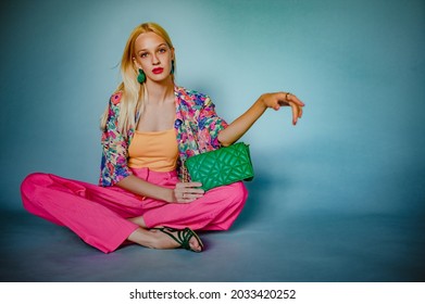 Fashinable woman wearing trendy summer outfit with colorful blouse, top, pink wide pants, strappy sandals, green quilted leather bag, posing, sitting on blue background. Copy, empty space for text