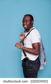 Fashion African Ethnicity Man Holding Denim Jacket On Back While Smiling At Camera And Standing On Blue Background. Cheerful And Handsome Young Person Wearing Pulling Suspenders. Studio Shot