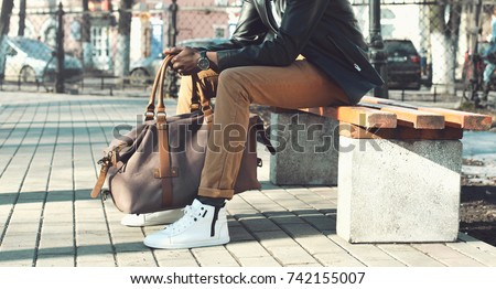 Fashion african man holding bag sitting on  bench in city park close up, male accessory 