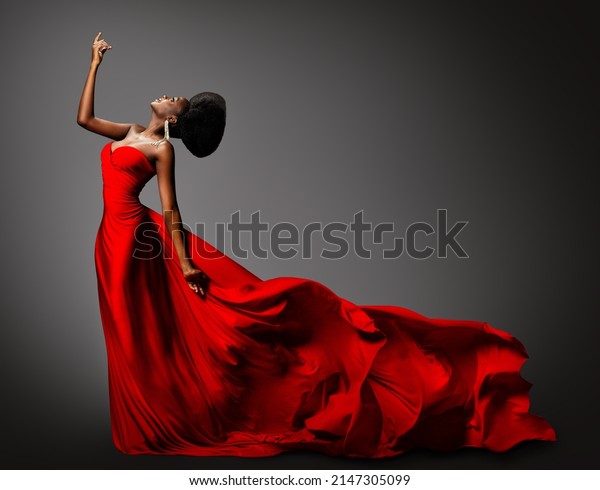 Fashion African Woman in Silk Dress dancing. Dark\
Skinned Model with Black Afro Hair in Long Evening Red Gown with\
Tail Fabric flying over Gray Background. Stylish Women Luxury\
Clothing Side View