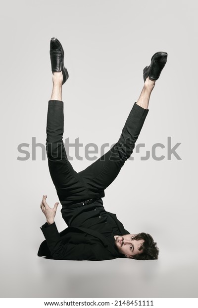 Fashion and Art. A stylish\
brunet man fashion model in an elegant black suit poses on a white\
background upside down with his legs up. Full-length studio\
portrait.