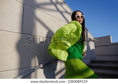 Fashion asian female model outdoor. Green down jacket, green skirt, green sweater, sunglasses. Monochromatic look. Urban city streets.